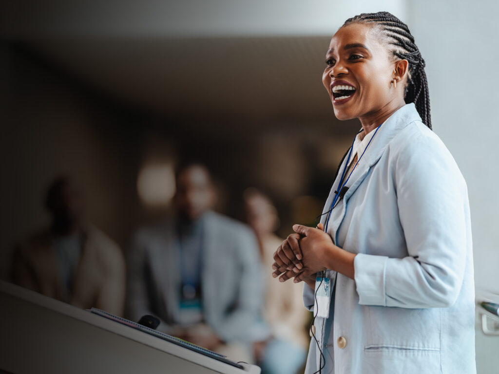 Professional African woman with a radiant smile engages attendees as she speaks at a business conference. With a blurred team in the backdrop, she embodies leadership and confidence.